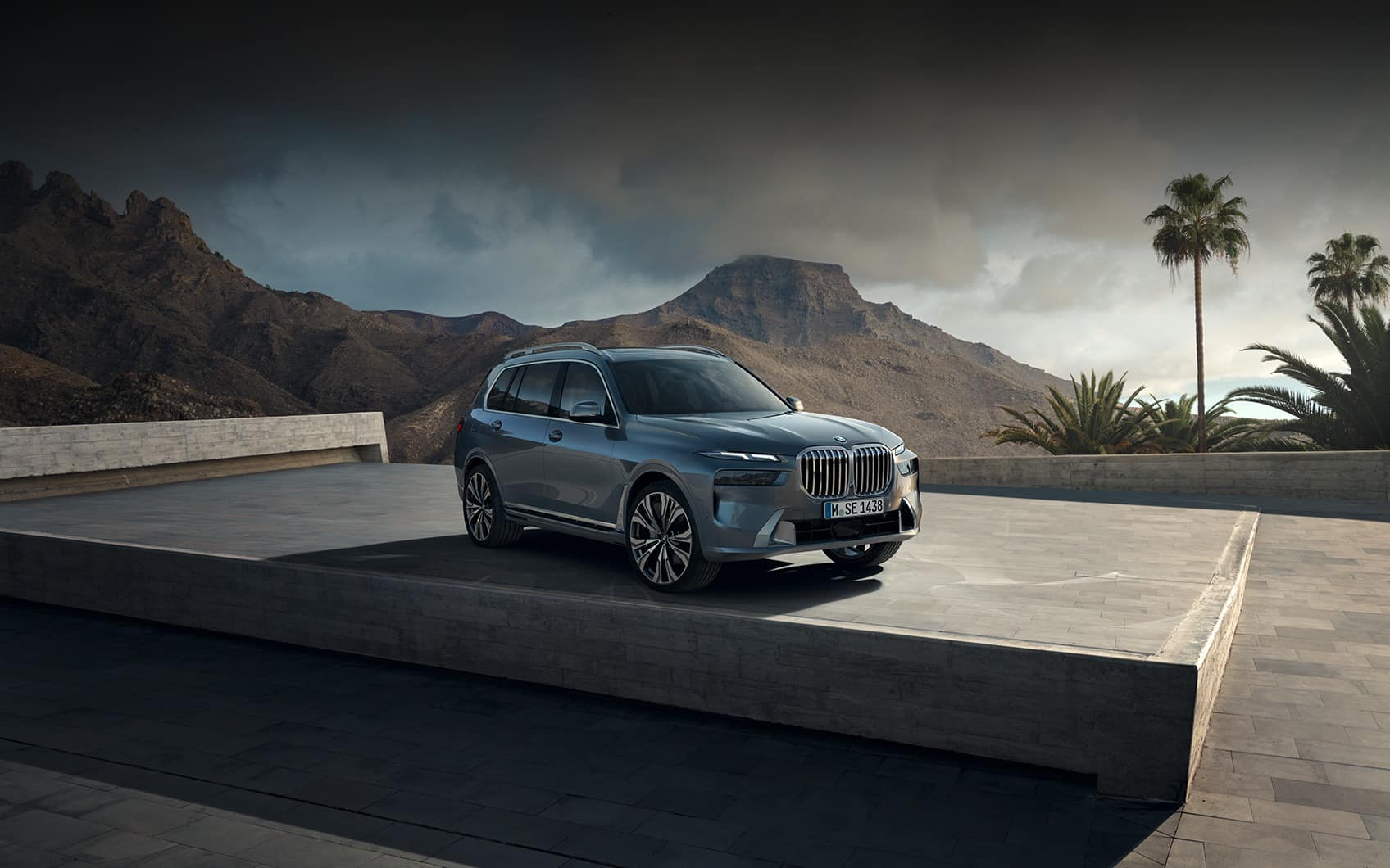 Silberner BMW X7 mit Y-Speichen-Felgen vor Bergkulisse und Palmen auf Betonplateau.