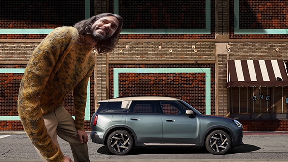 Grüner MINI Countryman Electric in Stadtumgebung mit modernen Gebäuden, kennzeichnendem Kühlergrill und Scheinwerfer