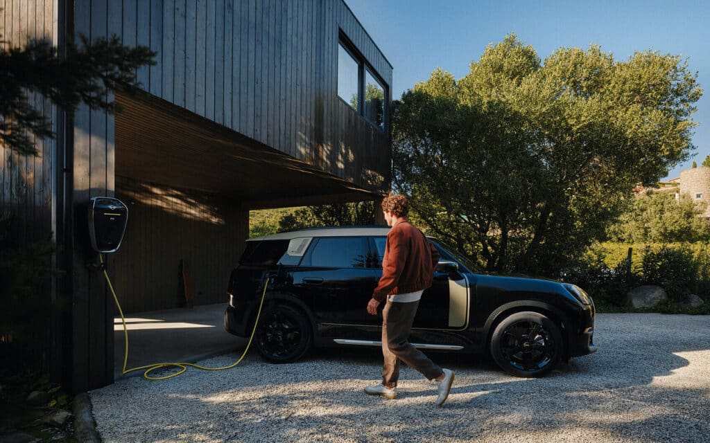 MINI Countryman Electric angeschlossen zum Laden an einer Wallbox vor einem modernen Wohnhaus mit Holzfassade