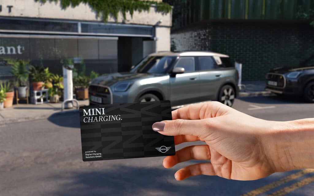 In das Bild hineinragende Hand hält eine MINI Charging Card vor ein MINI Modell mit urbaner Kulisse im Hintergrund