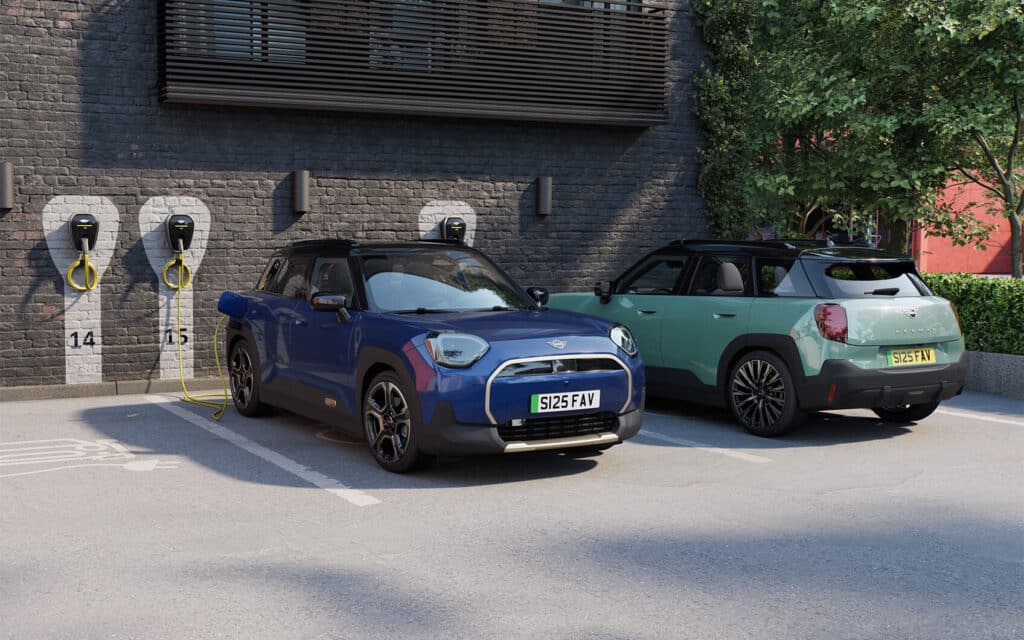 MINI Aceman und MINI Cooper Electric auf zwei Parkflächen vor einem Gebäude, jeweils angeschlossen zum Laden an eine Wallbox