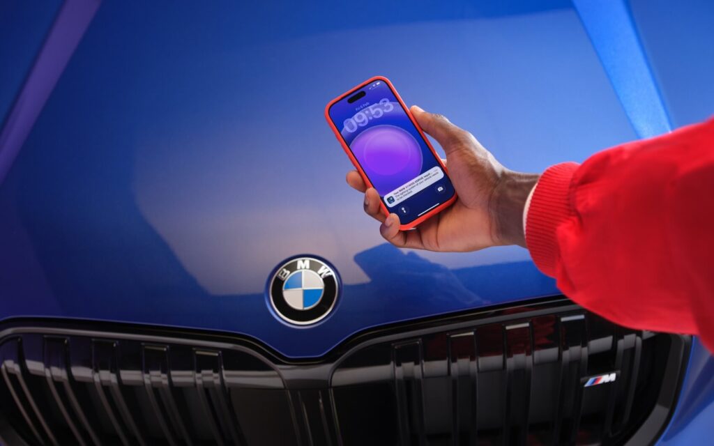Hand hält Smartphone mit BMW-Benachrichtigung vor der Front eines blauen BMW.