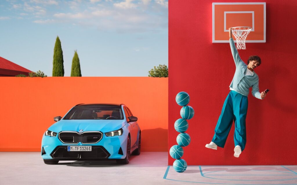 Mann hängt am Basketballkorb und schaut auf sein Smartphone neben blauem BMW und gestapelten Basketbällen.