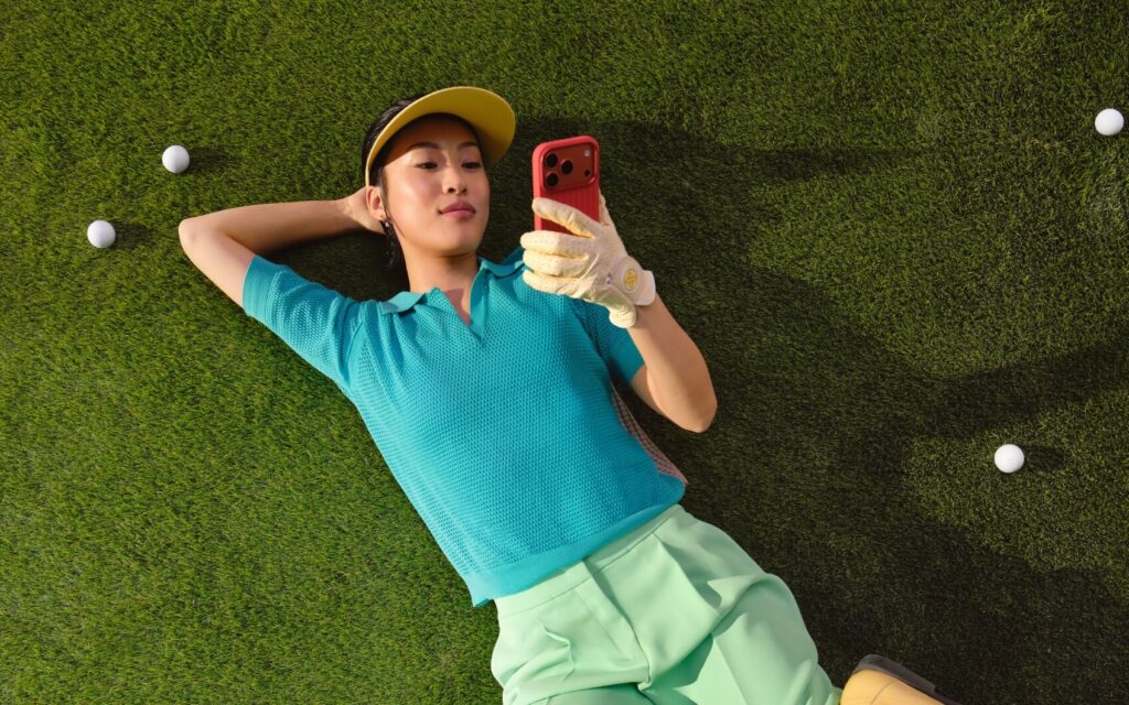 Golferin liegt auf dem Rasen und schaut auf ihr Smartphone, umgeben von Golfbällen
