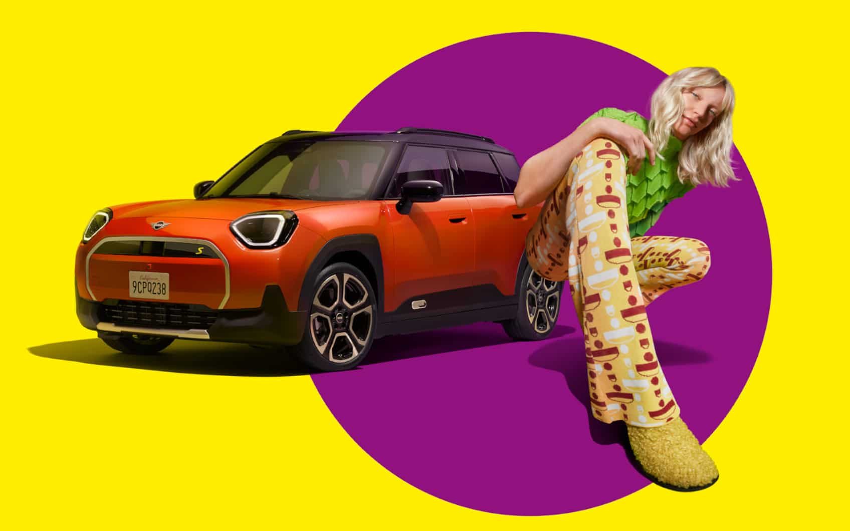 Oranger MINI Aceman vor gelbem Hintergrund mit violettem Kreis, neben dem MINI Aceman hockt eine Frau in bunter Kleidung